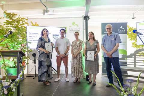 : Sisätilassa yleisön edessä seisoo viisi ihmistä lavalla: yksi on puhujapöydän luona ja toinen pitää edessään palkintotodistusta. Taustalla on kaksi näyttöä, jossa lukee Hatsinanpuisto park ja palkintoa vastaanottavien henkilöiden nimet: Pia Kuusiniemi, Principal Designer, Milla Hakari, Partner, Felix Bourgeau Project Designer, Loci Landscape architects ja Kristina Rocha City of Espoo. 