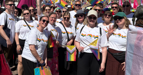 City of Espoo employees participating in the Pride parade are looking at the camera. A large group of other participants can be seen in the background.