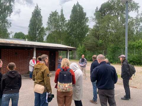 Ihmiset seisovat ringissä kuuntelemassa yhtä henkilöä. Taustalla matala tiilirakennus, hiekkakenttä ja vihreitä puita.