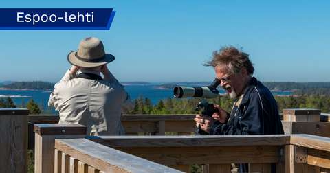 Two people in a birdwatching tower. One has their back to the camera and is looking out to sea through binoculars, while the other is looking through a spotting scope.