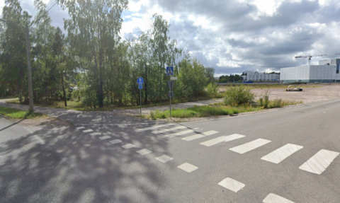 Etualalla katu sekä puita ja pensaita, takana iso hiekkakenttä, joka on tulevaa rakentamisaluetta.