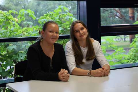 Two students are sitting next to each other at a table. A green forest can be seen in the background.