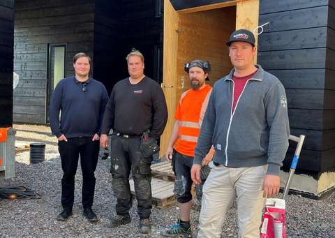 Fyra personer framför en svart träbyggnad.