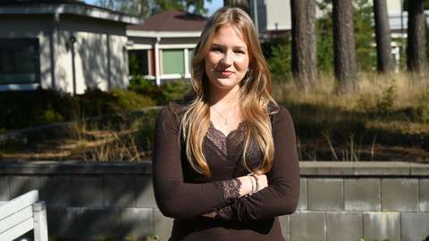 A student is standing outdoors with their arms crossed. Pine trees can be seen in the background.