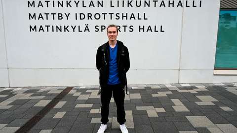 A student is standing outdoors in front of a wall displaying Matinkylän liikuntahalli in three languages.