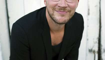 Mustiin pukeutunut kalju mies nojaa käsillä reisiin ja katsoo hymyillen kohti.