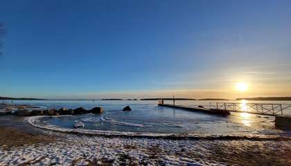 Kuvassa näkyy laituri, jonka takana näkyy auringonlasku. Etualalla on rannan hiekkaa ohuen lumipeitteen alla ja veden pinnalla on jäätä.