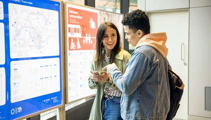 Kaksi nuorta seisovat joukkoliikenteen aikataulujen edessä ja tutkivat kännykästä reittivaihtoehtoja.