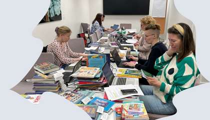 Opettajia ja kirjastolaisia työskentelemässä pitkän pöydän ääressä. Pöydällä on pinoissa paljon uusia lasten- ja nuortenkirjoja.