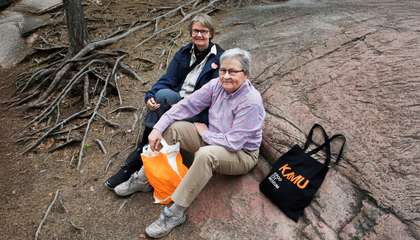 Sateenkaarisenioreiden Anne ja Päivi istuvat kalliolla ja katsovat ylös kameraan.