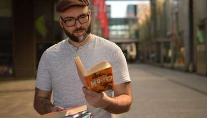 An adult is standing outside reading a book. In addition to the book they are reading, the person is carrying other books.