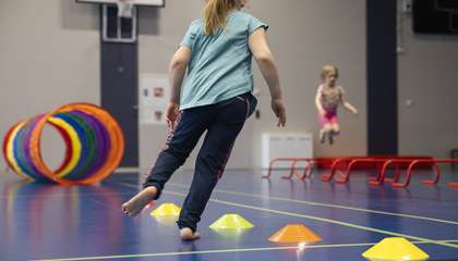 Kuvassa lapsi pujottelee juosten liikuntasalin lattialle laitettujen tötsien välistä. Taustalla näkyy toinen lapsi hyppäämässä esteen yli.
