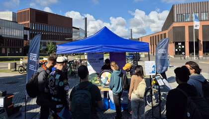 Pyöräilyä edistävä telttapiste Aalto-yliopiston aukiolla. Teltan eteen on kerääntynyt useita opiskelijoita, ja yksi opiskelijoista on pyöräyttämässä onnenpyörää.