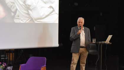 Luennoitsija seisoo auditorion lavalla ja puhuu mikrofoniin.