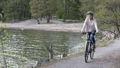 Kuvassa nainen ajaa pyörällä hiekkatiellä veden äärellä. Takana hiekkarantaa ja puita.