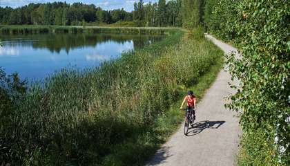 En cyklist på en sandväg vid en tjärn, i bakgrunden en skogsdunge.