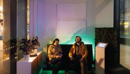 Kaksi ihmistä istuvat penkillä tunnelmallisesti valaistussa hämärässä odotustilassa. Molemmilla on päässä mustat kuulokkeet, ja heidän taustallaan seinällä on kaksi valkoista akustiikkapaneelia.