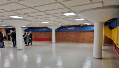 Iso, matala ja tyhjä hallimainen tila, jossa seinät tiiltä ja osittain  maalattu eri väreillä. Kolme iso pylvästä, yhden takana seisoo joukko ihmisiä.