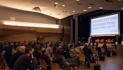 Isossa salissa ihmisiä istuu tuoliriveillä kuuntelemassa puhujaa lavalla. Puhujan taakse lavalle on heijastettu esitys.